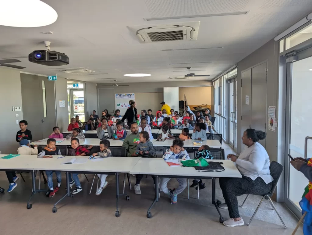 Children and parents seated at long tables during an ELSP language class in a community hall